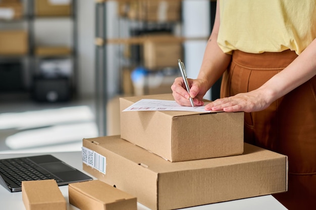 Closeup of young female worker of warehouse writing down personal data on consignment on top of stack of boxes in front of camera