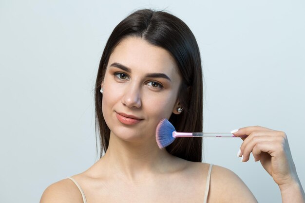 Closeup of a young girl in a light top on a white background making a facial makeup A pretty woman holds a cosmetic brush near her face and smiles