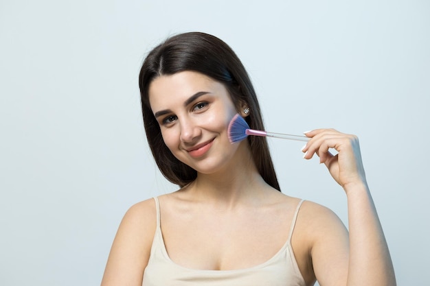 Closeup of a young girl in a light top on a white background making a facial makeup A pretty woman holds a cosmetic brush near her face and smiles