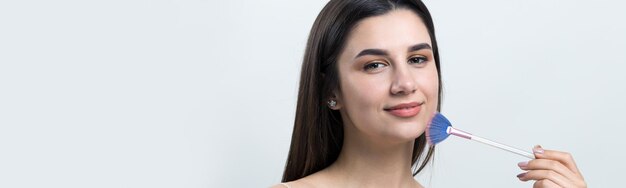 Closeup of a young girl in a light top on a white background making a facial makeup A pretty woman holds a cosmetic brush near her face and smiles