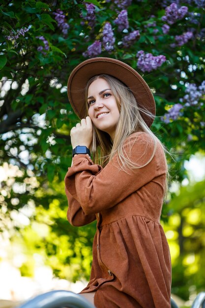 Closeup young girl standing on a background of lilac bushes. she is of European appearance in a brown hat and dress