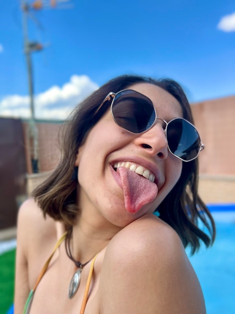 Closeup of a young girl in sunglasses sticking out her tongue at camera while enjoying in the pool at home in summer