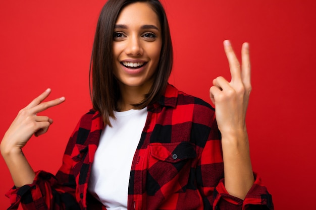 Foto primo piano di giovane donna bella bruna fresca e positiva felice con emozioni sincere che indossa t-shirt bianca e camicia a quadri rossa alla moda isolata su sfondo rosso con spazio libero e divertirsi