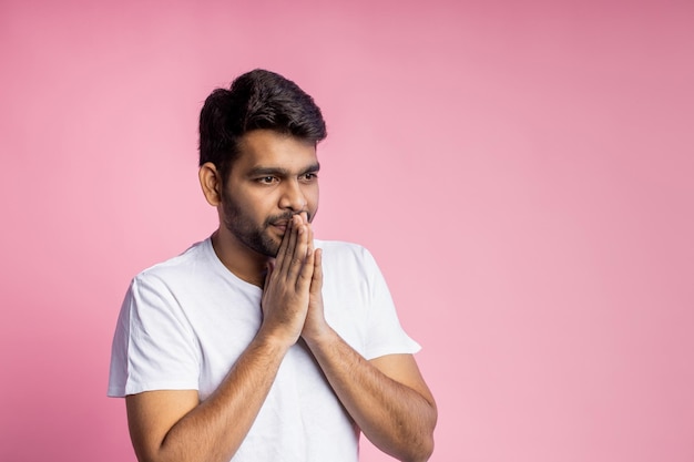 Closeup of young indian male in white t shirt, praying with palms clasped together, asking the God for good luck, success, health, on pink background with copy space. Human emotion, belief concept.