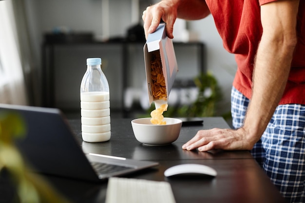 Primo piano del giovane che mette i fiocchi di mais in una ciotola per farli con il latte per la colazione a tavola con l