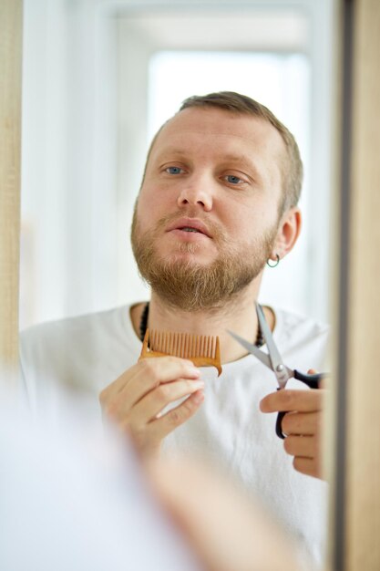 Photo closeup of a young man trimming his beard with scissors indoors