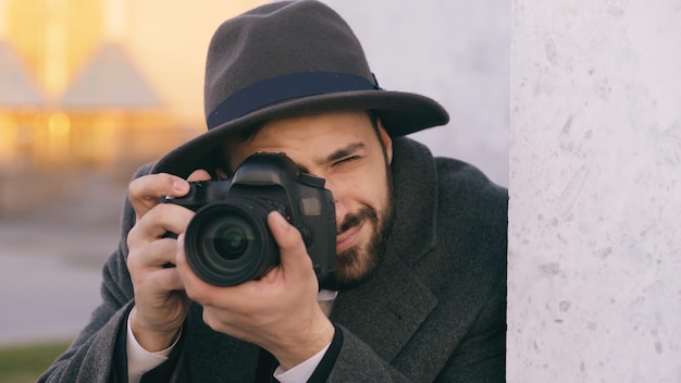 壁の後ろでスパイしながらカメラで有名人を撮影する帽子の若いpaparazzi男のクローズアップ