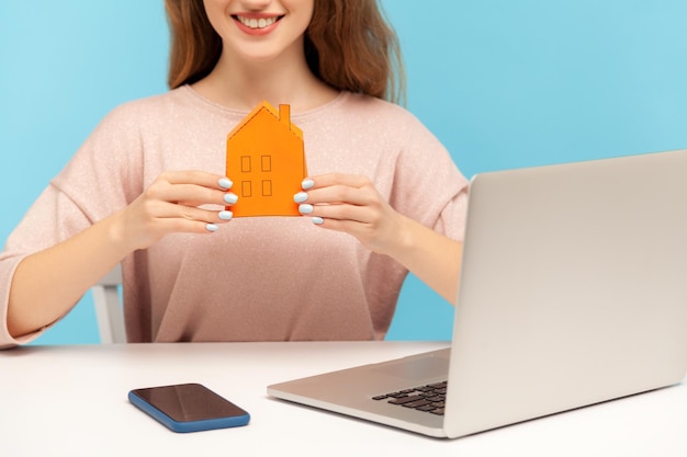 Closeup of young positive woman, real estate agent sitting at workplace with laptop and holding small paper house, smiling to camera, offering rental and home purchase. indoor studio shot isolated