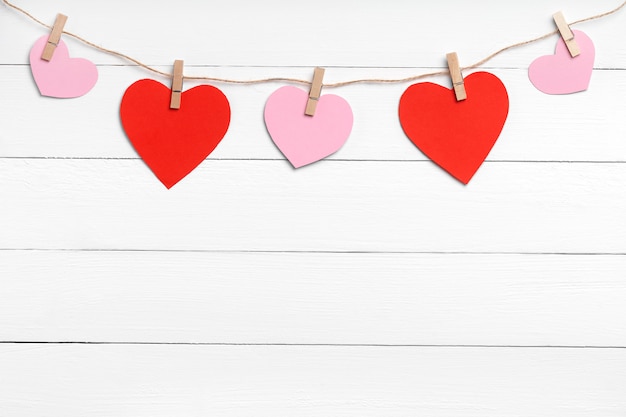 Clothespins with colored hearts hanging on rope behind white wooden surface. Copy space, top view.