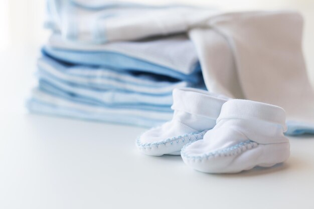 clothing, babyhood, motherhood and object concept - close up of white baby bootees with pile of clothes for newborn boy