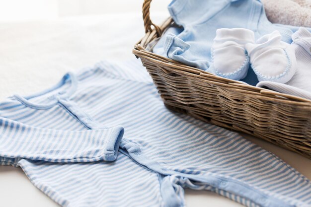 clothing, babyhood, motherhood and object concept - close up of white baby bootees with pile of clothes and towel for newborn boy in basket on table