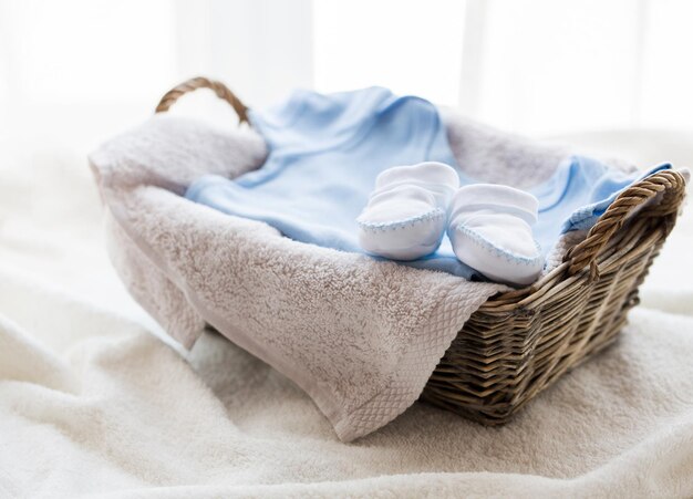 clothing, babyhood, motherhood and object concept - close up of white baby bootees with pile of clothes and towel for newborn boy in basket on table