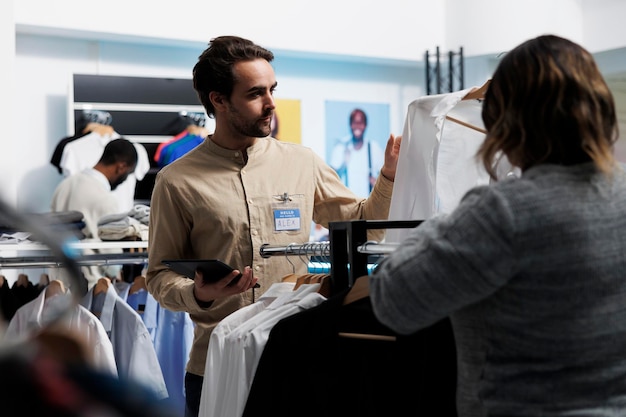 衣料品店のクライアントがシャツをハンガーに掛け、コンサルタントにサイズのオプションについて尋ねます。デジタル タブレットを保持し、顧客を支援するショッピング センターのファッション ブティックの従業員