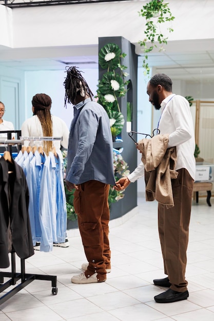 Clothing store employee examining shirt on customer and checking fit. African american man choosing menswear in shopping mall while getting help from fashion boutique assistant