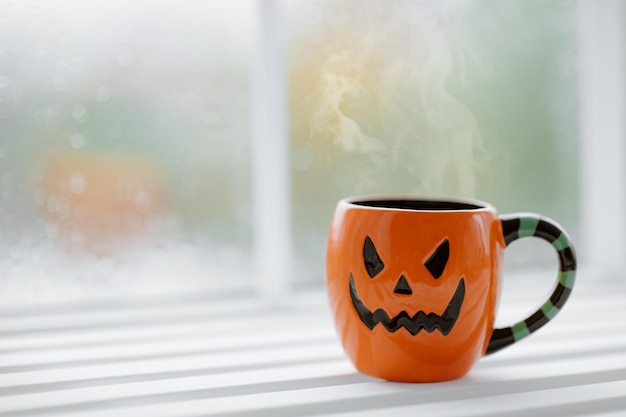 Clouse up of cup in shape of pumpkin Jack O lantern with hot coffee on windowsill