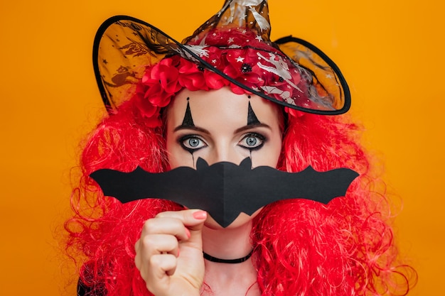 Clown girl in halloween costume covers her face with paper bat isolated on orange