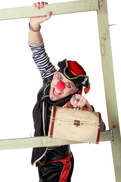 Клоун одет в костюм пирата и держит в руке сундук с сокровищами на белом фоне