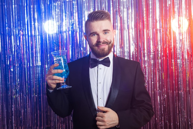 Clubfeest en vakantie concept - portret van elegante knappe man in een duur pak houdt blauwe cocktail vast op glanzende achtergrond.