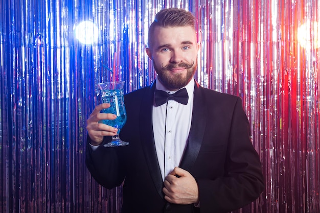 Clubfeest en vakantie concept - portret van elegante knappe man in een duur pak houdt blauwe cocktail vast op glanzende achtergrond.