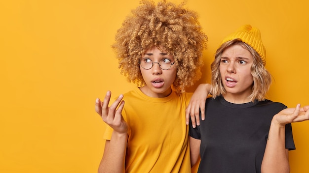 Foto clueless aarzelende jonge vrouwen voelen zich beschaamd en ondervraagd, halen hun schouders op omdat ze zich niet bewust zijn van wat er gebeurt, gekleed in casual t-shirts, gefocust weg fronsende gezichten geïsoleerd over gele achtergrond