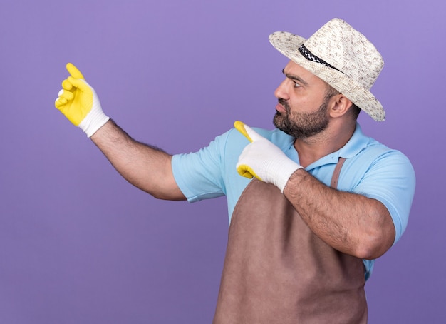 clueless adult male gardener wearing gardening hat and gloves looking and pointing at side isolated on purple wall