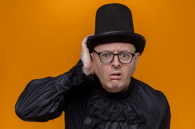 Clueless adult man with top hat and glasses in black gothic shirt putting hand on his hat and looking 