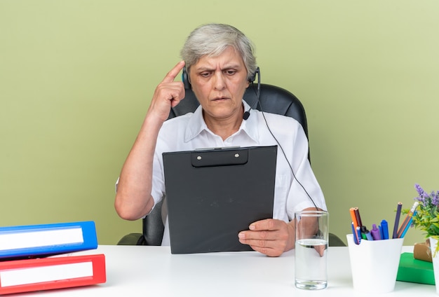Clueless Kaukasische vrouwelijke callcenter operator op koptelefoon zittend aan een bureau met office tools houden en kijken naar klembord geïsoleerd op groene muur