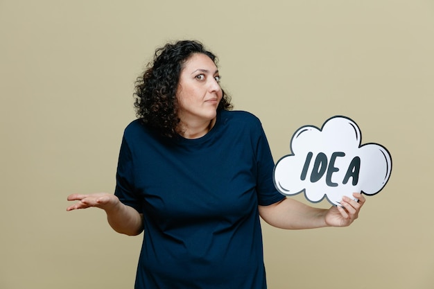 Clueless middleaged woman wearing tshirt showing idea bubble and empty hand looking at camera isolated on olive green background