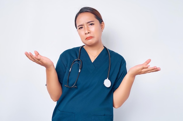 Clueless young Asian woman nurse wearing blue uniform with a stethoscope shrugging and spread hands sideways unaware dont now cant help isolated on white background Healthcare medicine concept