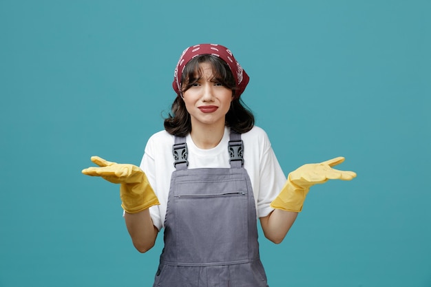 Clueless young female cleaner wearing uniform bandana and rubber gloves looking at camera showing empty hands isolated on blue background