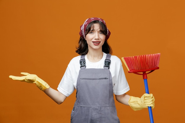 Clueless young female cleaner wearing uniform rubber gloves and bandana holding squeegee mop looking at camera showing empty hand isolated on orange background
