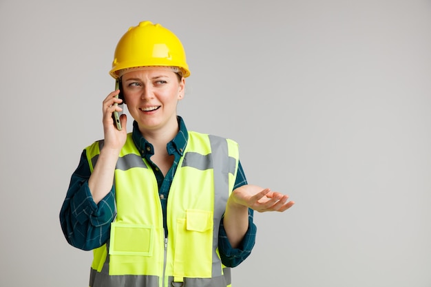 Clueless young female construction worker wearing safety helmet and safety vest talking on phone looking at side showing empty hand 