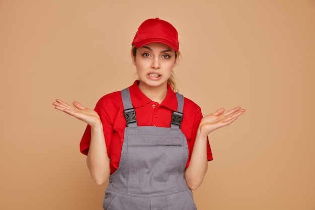 Clueless young female construction worker wearing uniform and cap showing empty hands 