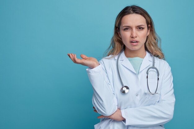 Clueless young female doctor wearing medical robe and stethoscope around neck showing empty hand 