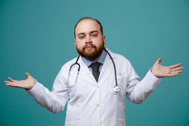 Giovane medico di sesso maschile senza indizi che indossa un camice medico e uno stetoscopio intorno al collo che mostra le mani vuote guardando la fotocamera isolata su sfondo blu