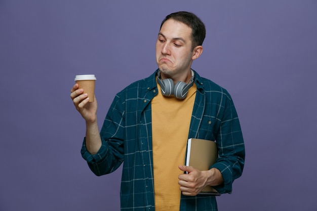 Clueless young male student wearing headphones around neck holding note book under arm holding paper coffee cup looking at it isolated on purple background