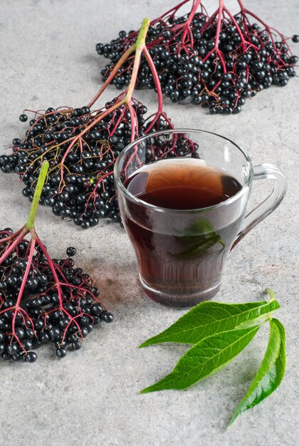 Grappoli di sambuco con foglie verdi e bevanda in un bicchiere su sfondo grigio.