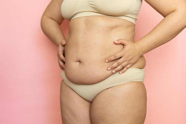 Photo clutch woman sagging belly closeup folds on stomach loose skin and cellulite obesity naked overweight plus size girl on pink background in beige underwear concept of dieting and body control