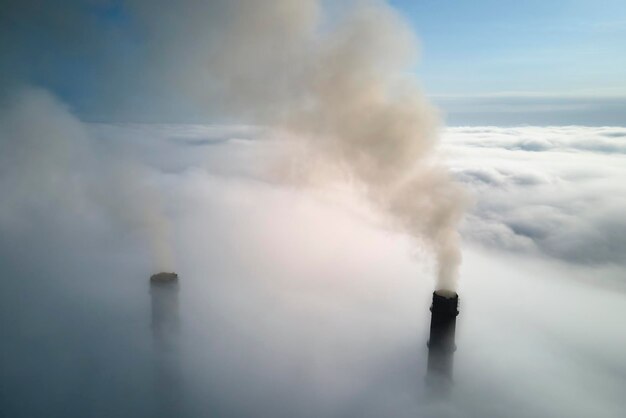 Coal power plant high pipes emitting black poisonous smoke moving upwards over white fog polluting air Production of electrical energy with fossil fuel concept