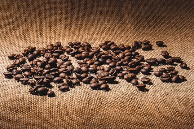 Coarse coffee beans, roasted and not ground, are scattered on a textured fabric with a burlap in warm colors.