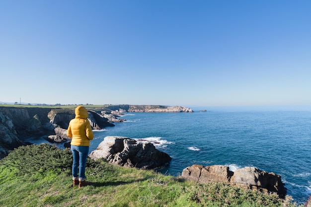 Paesaggio costiero e ragazza in cappotto giallo con cappuccio guardando il mare e camminando copia spazio