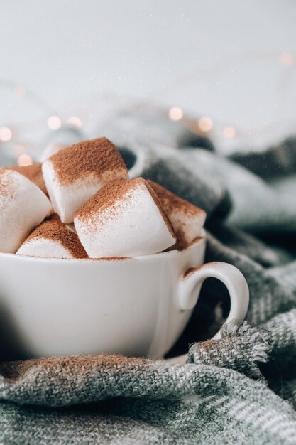 Cocoa drink with marshmallow and cacao powder on cozy winter sweater background. Christmas lights. Winter holiday home atmosphere