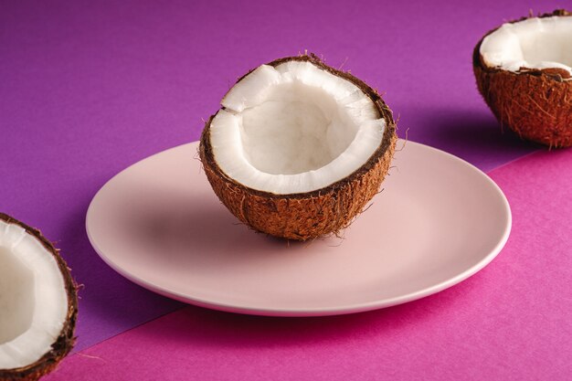 Coconut half in pink plate with nut fruits on violet and purple plain surface