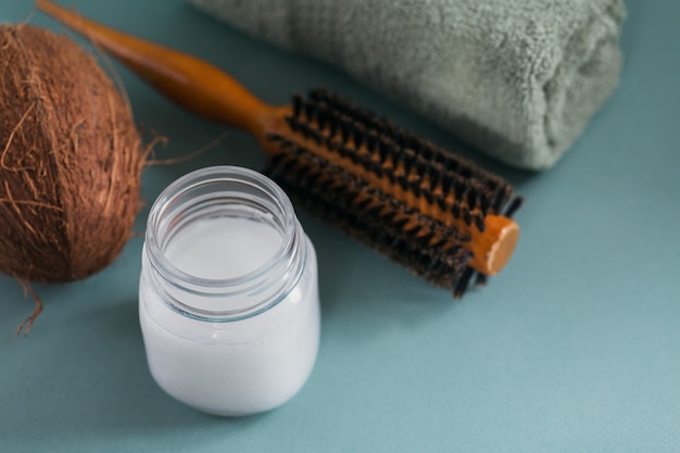 Coconut oil in a bottle with coconuts, towel and hairbrush on light blue surface