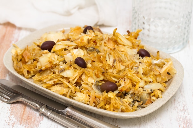 Codfish with potato chips and olives on plate on white surface