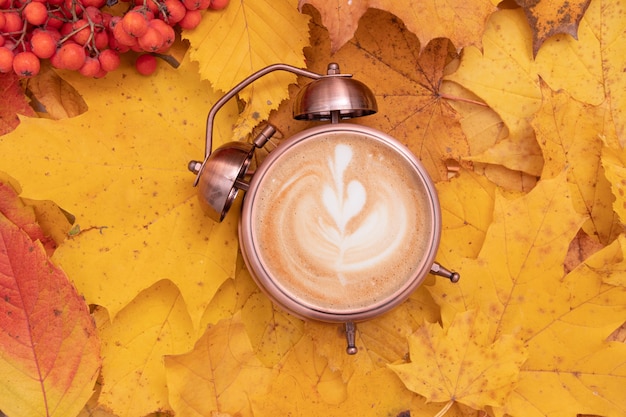 Искусство кофе в будильнике на фоне опавших листьев. Осеннее время и концепция утреннего кофе