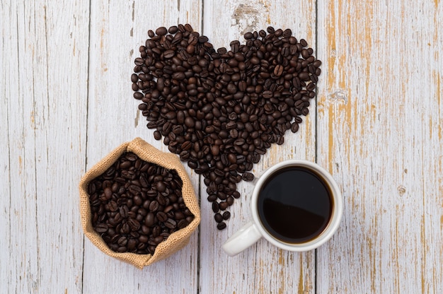 Coffee beans arranged in a heart shape, of coffee and a coffee mug on light wood background