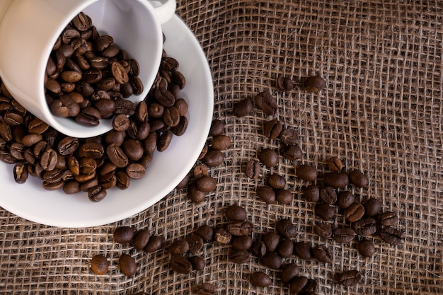 Coffee beans scattered from a porcelain white cup on burlap