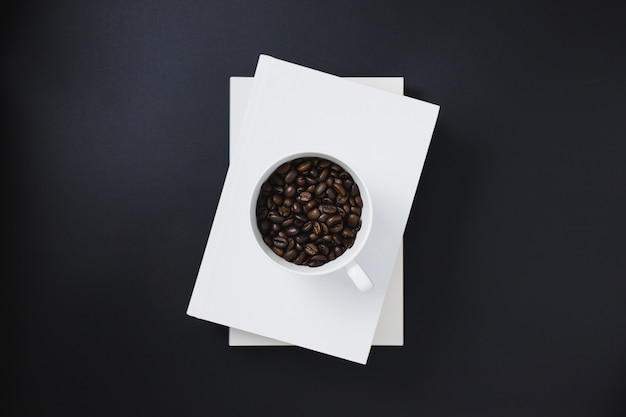 Coffee beans in a white coffee mug placed on white books stacked on a black background