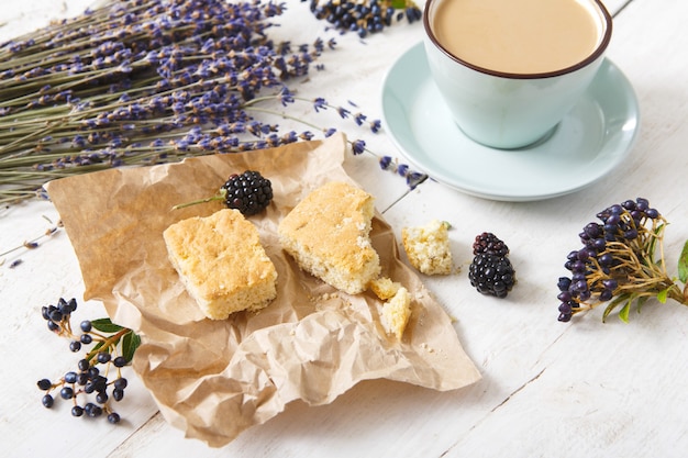 Coffee, biscuits, berries and lavender flowers composition, closeup on white wood. Blue cup with creamy foam